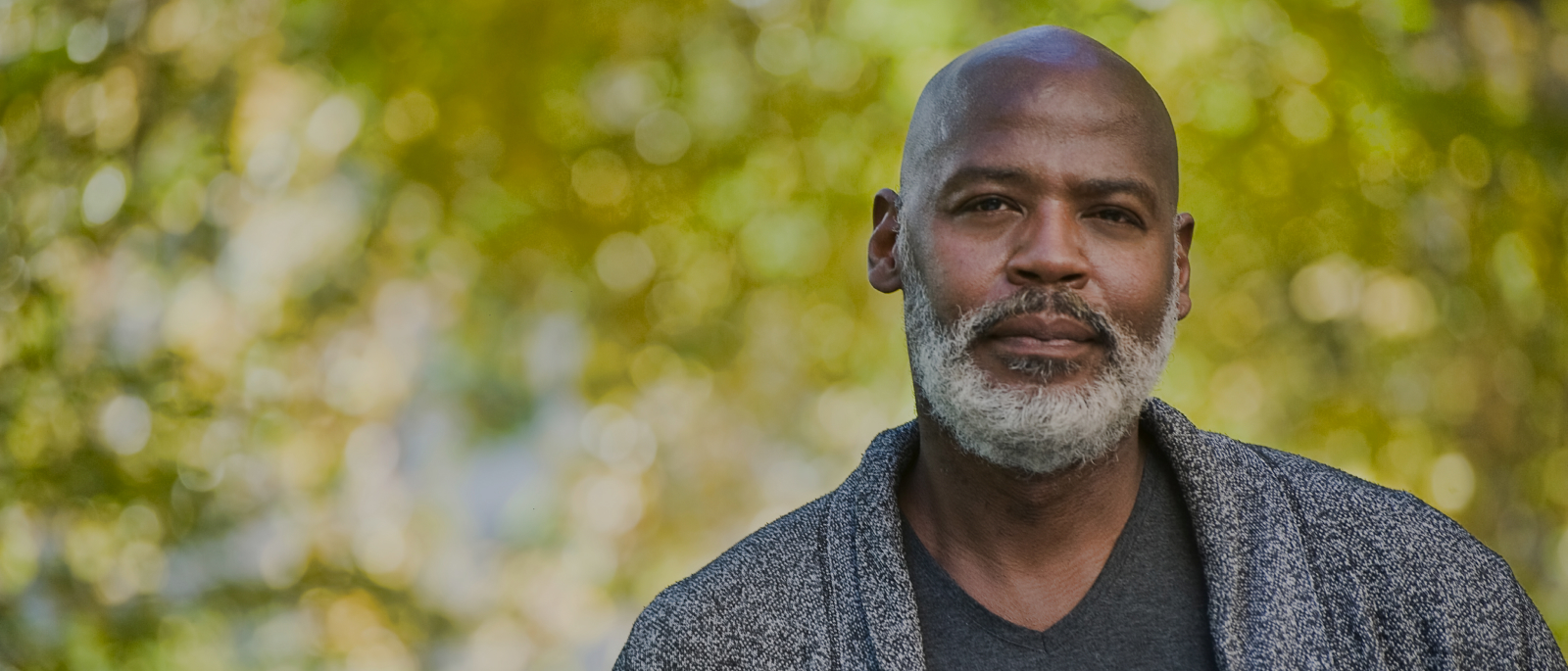A black man with a gray beard thoughtfully looks toward the camera.  Behind him are green trees.